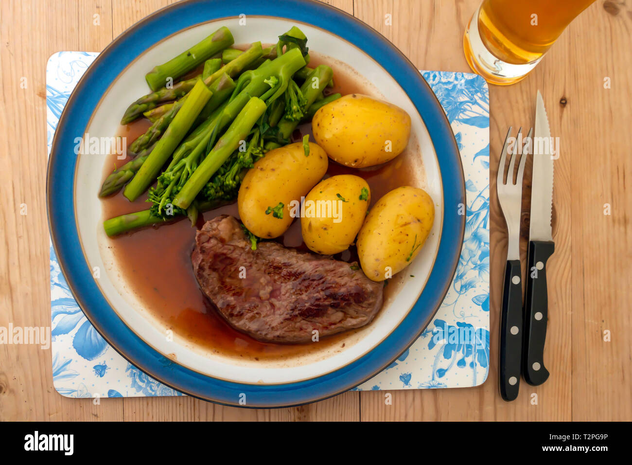 Un fritto Aberdeen Angus Scamone bistecca di manzo servita con patate di primizia, filetto di broccoli e asparagi e sugo di carne su un blu orlato piastra su una tavola di legno Foto Stock