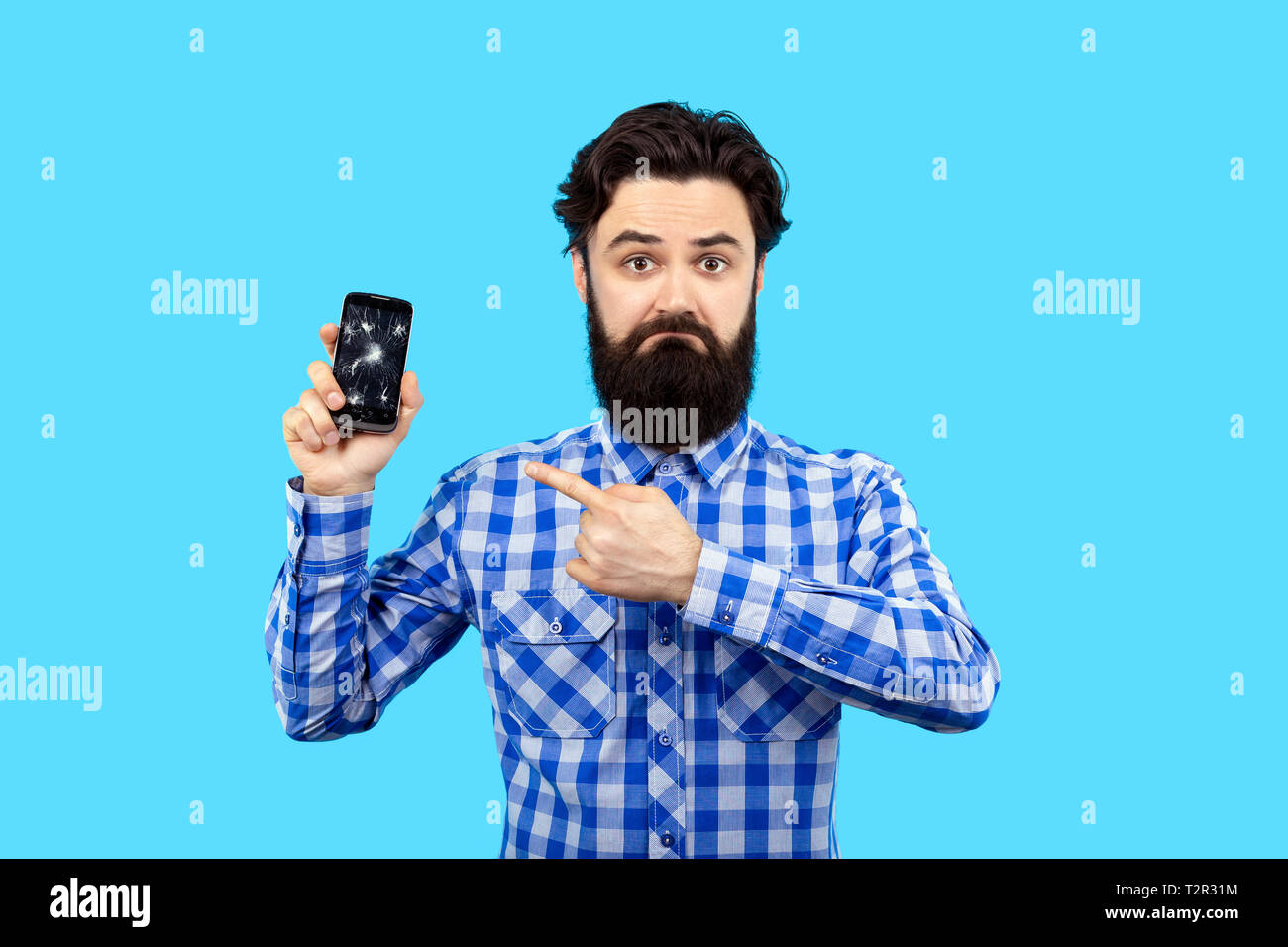 Barbuto triste hipster uomo puntare il dito a rotto lo schermo dello smartphone, immagine su blu bacground, concetto di ricerca servizio gsm Foto Stock