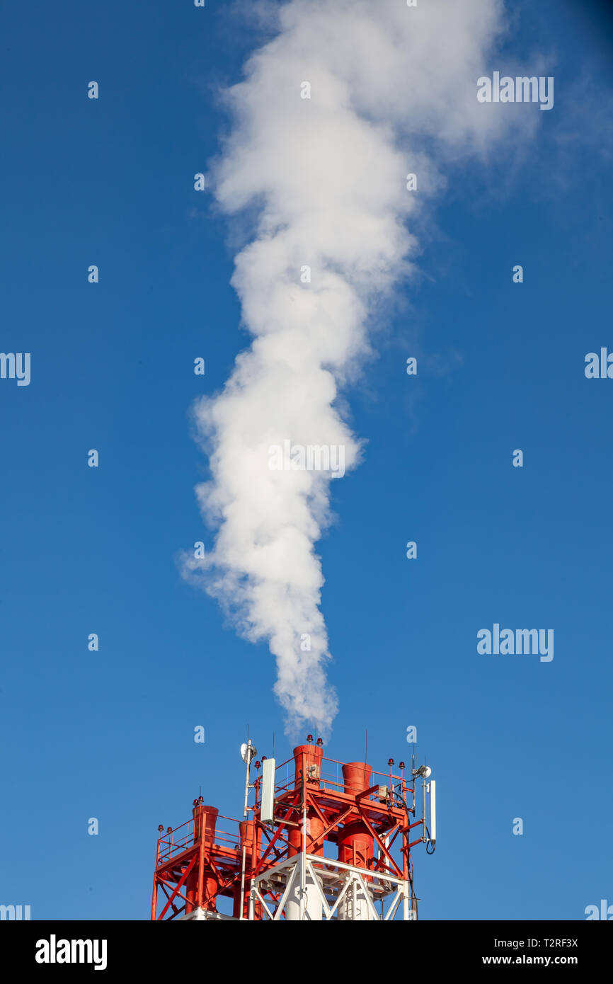 White nocivi del fumo proveniente al di fuori del rosso-e-bianco con tubi di comunicazione mobile antenne in una fabbrica nel centro della città contro lo sfondo di Foto Stock