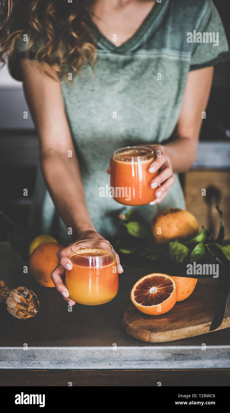 Giovani femmine tenendo due bicchieri di spremuta d'arancia rossa o frappè in mani nei pressi di calcestruzzo contatore da cucina. Uno stile di vita sano, vegan, Foto Stock