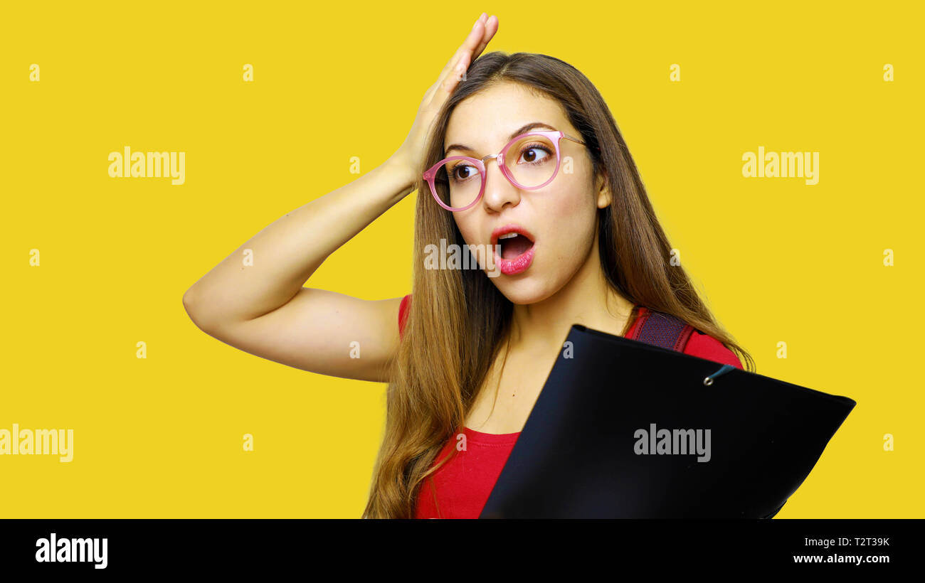 Spaventata giovane donna con la bocca aperta tenendo la testa con la mano e indossa gli occhiali e red t shirt porta cartella, dimentica di fare qualcosa di importante Foto Stock