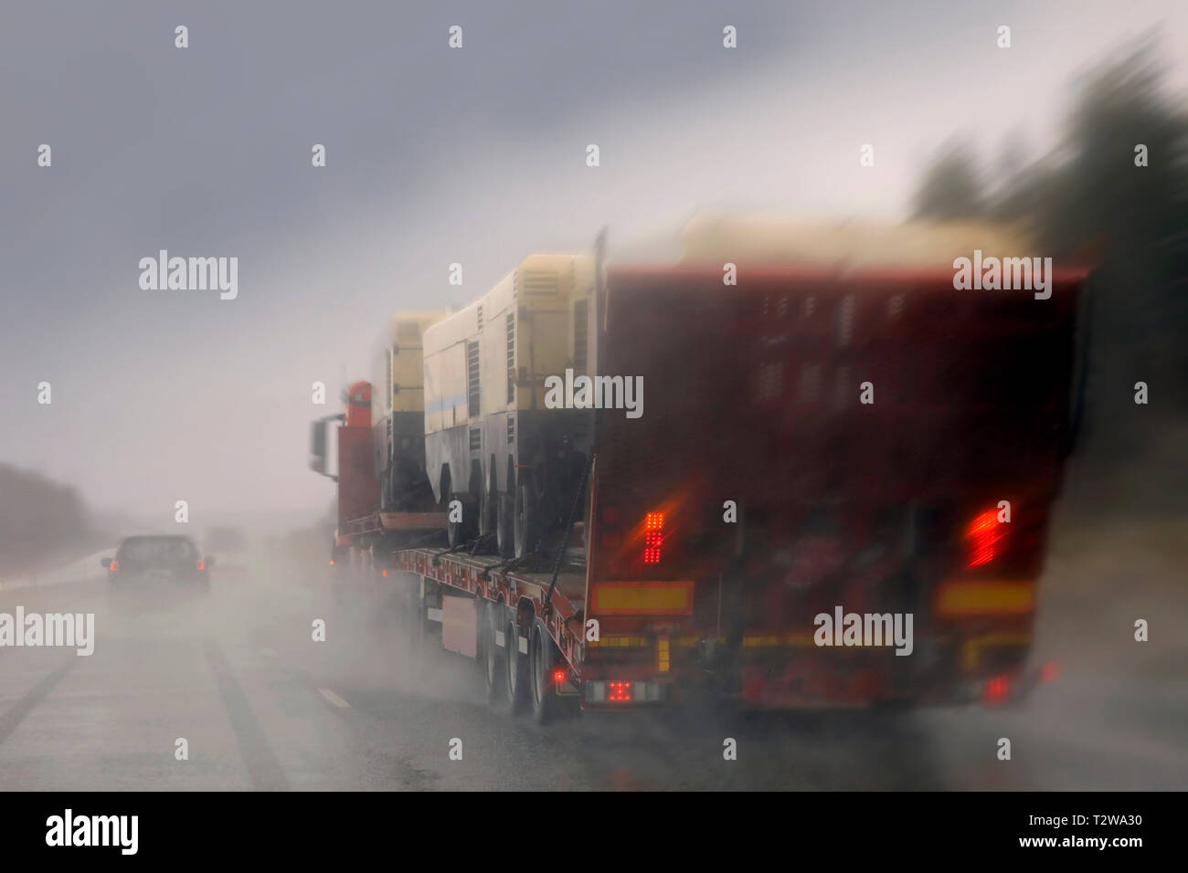 La guida lungo l'autostrada in pioggia e passando un semi carrello con un carico di macchinari, di spruzzi di acqua piovana, vista offuscata. Foto Stock