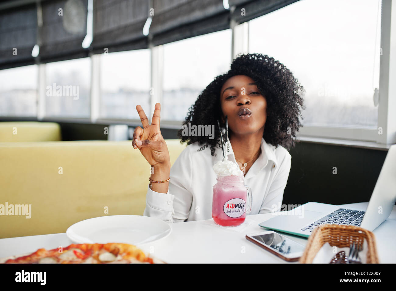 Сheerful business americano africano donna con capelli afro, usura camicetta bianca seduta a tavola, lavorare con il computer portatile del caffè e drink frullato rosa cocktai Foto Stock