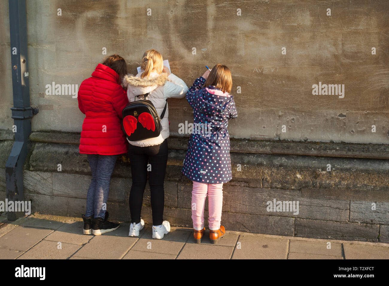 La scuola dei bambini in un giorno di viaggio a Oxford in Colorati luminosamente vestiti compila il loro tour risposte ai quiz appoggiata contro un antico muro di pietra Foto Stock