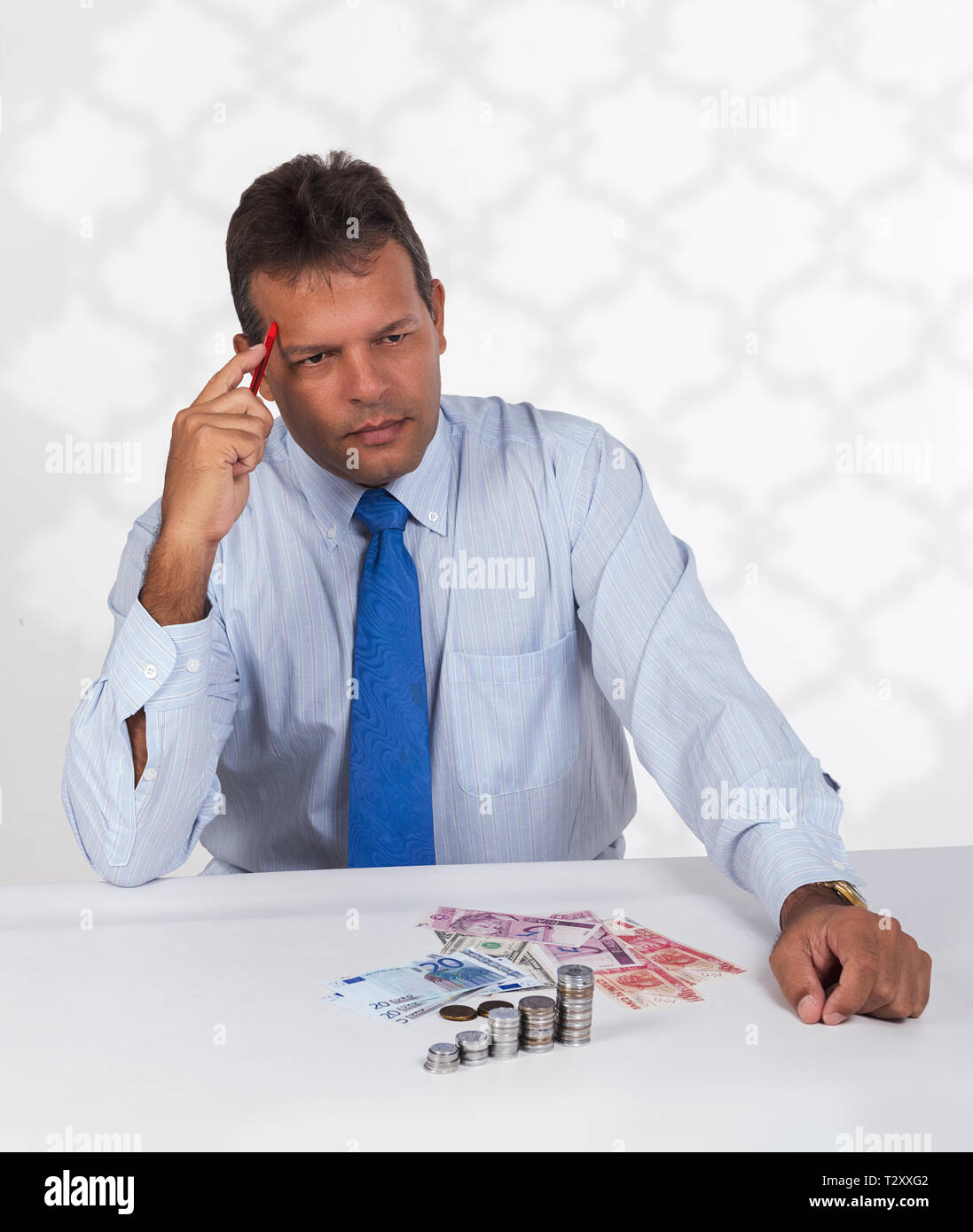 Un latino uomo seduto davanti a un gruppo di banconote e monete con un pensiero o espressione di calcolo sul suo volto Foto Stock