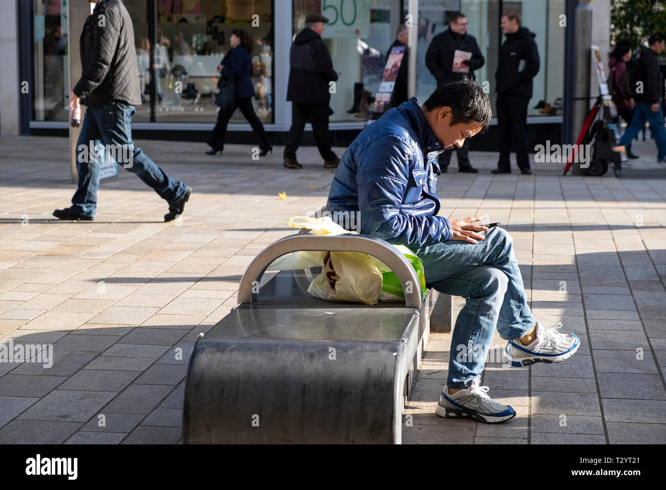 Giovane uomo in giacca blu e jeans seduti su una città banco di calcestruzzo controllando i messaggi sul suo telefono cellulare Foto Stock