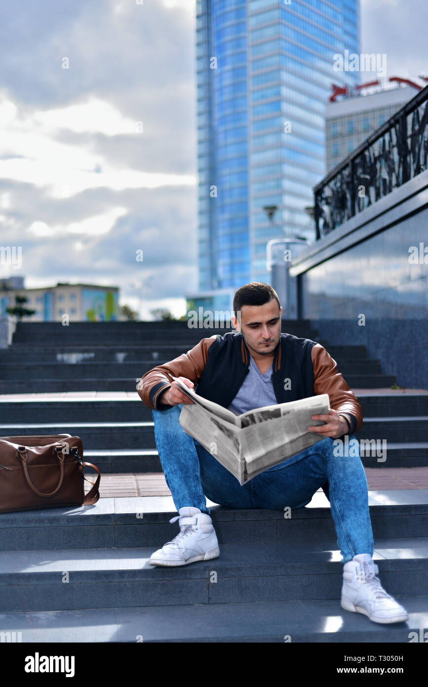 Ritratto di giovane ragazzo in giacca di pelle leggendo il giornale in città. l uomo è seduto sui gradini ofladder. successiva è di borsa. edifici alti in backgr Foto Stock