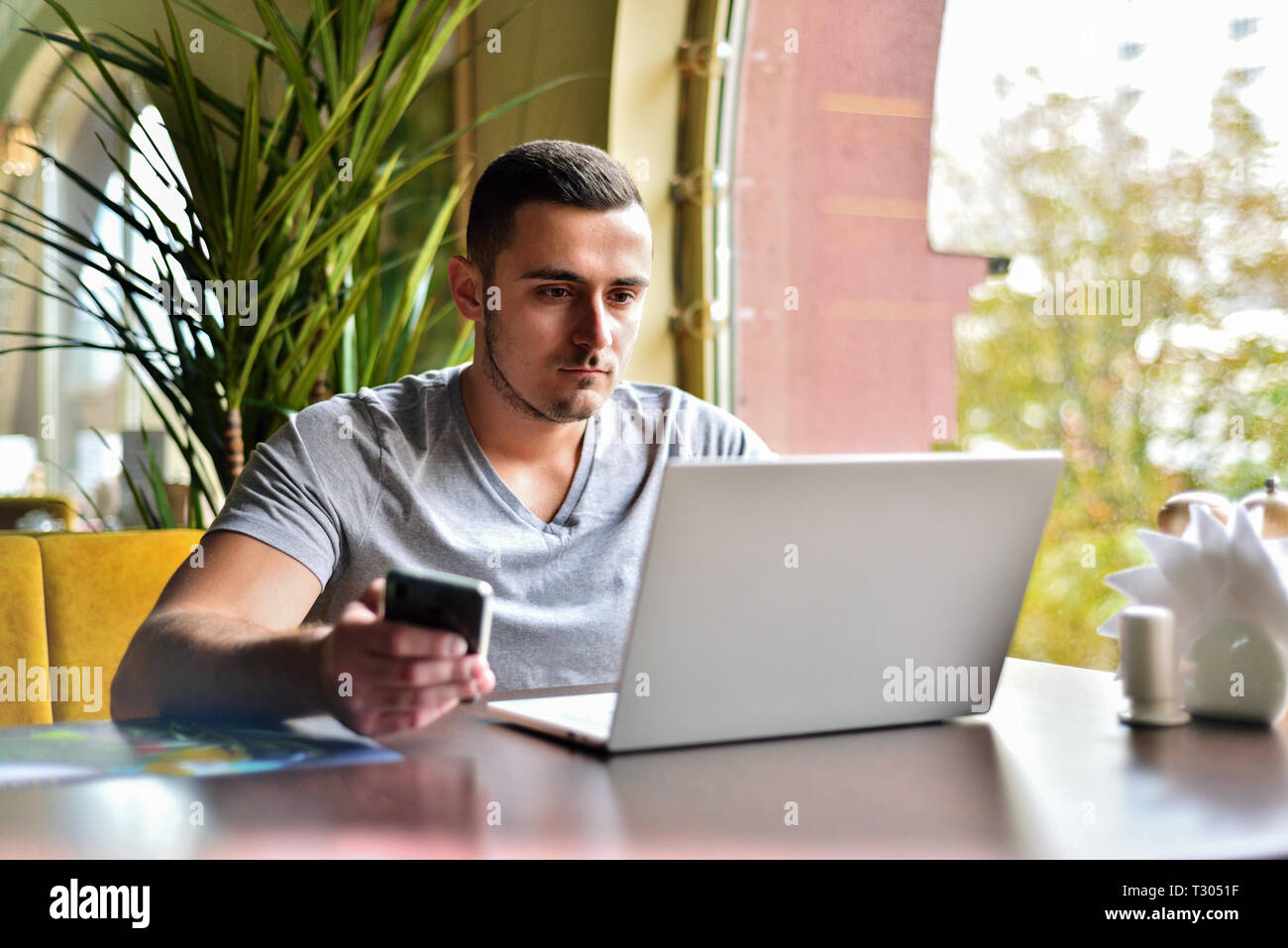 Giovane ragazzo è libero professionista in cafe lavorando dietro laptop. l uomo detiene il telefono in mano e invia il messaggio Foto Stock