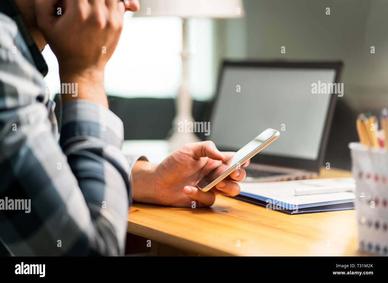 Lavoratore pigro usando il telefono in ufficio evitando il lavoro. Annoiato, triste o uomo infelice appoggiata contro la mano e la navigazione dei social media su Internet con lo smartphone. Foto Stock