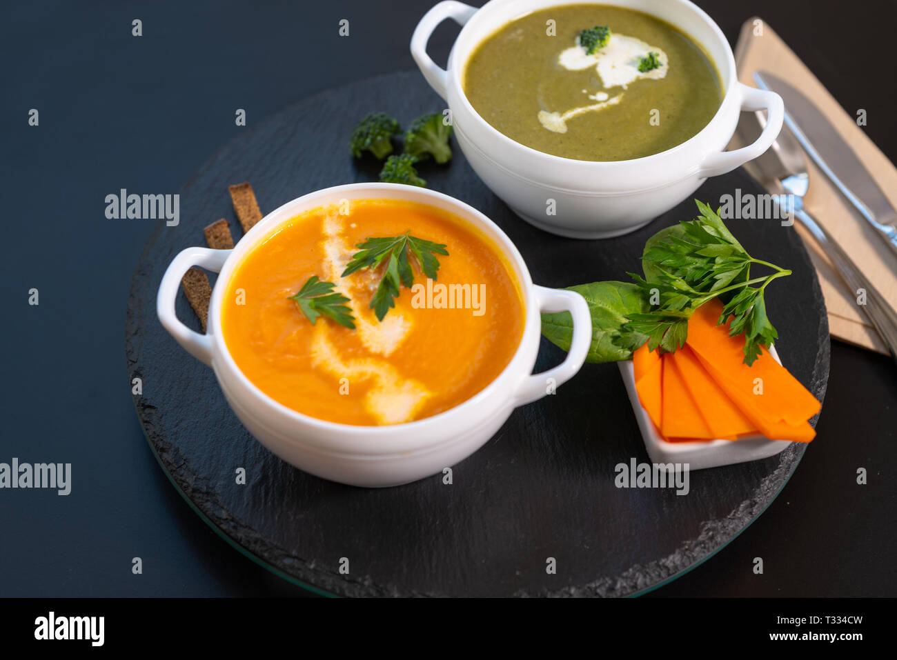 Due ciotole di zucca e broccoli minestra di crema di latte servita su una circolare in legno nero di bordo su una corrispondente sfondo nero con tovagliolo e posate Foto Stock