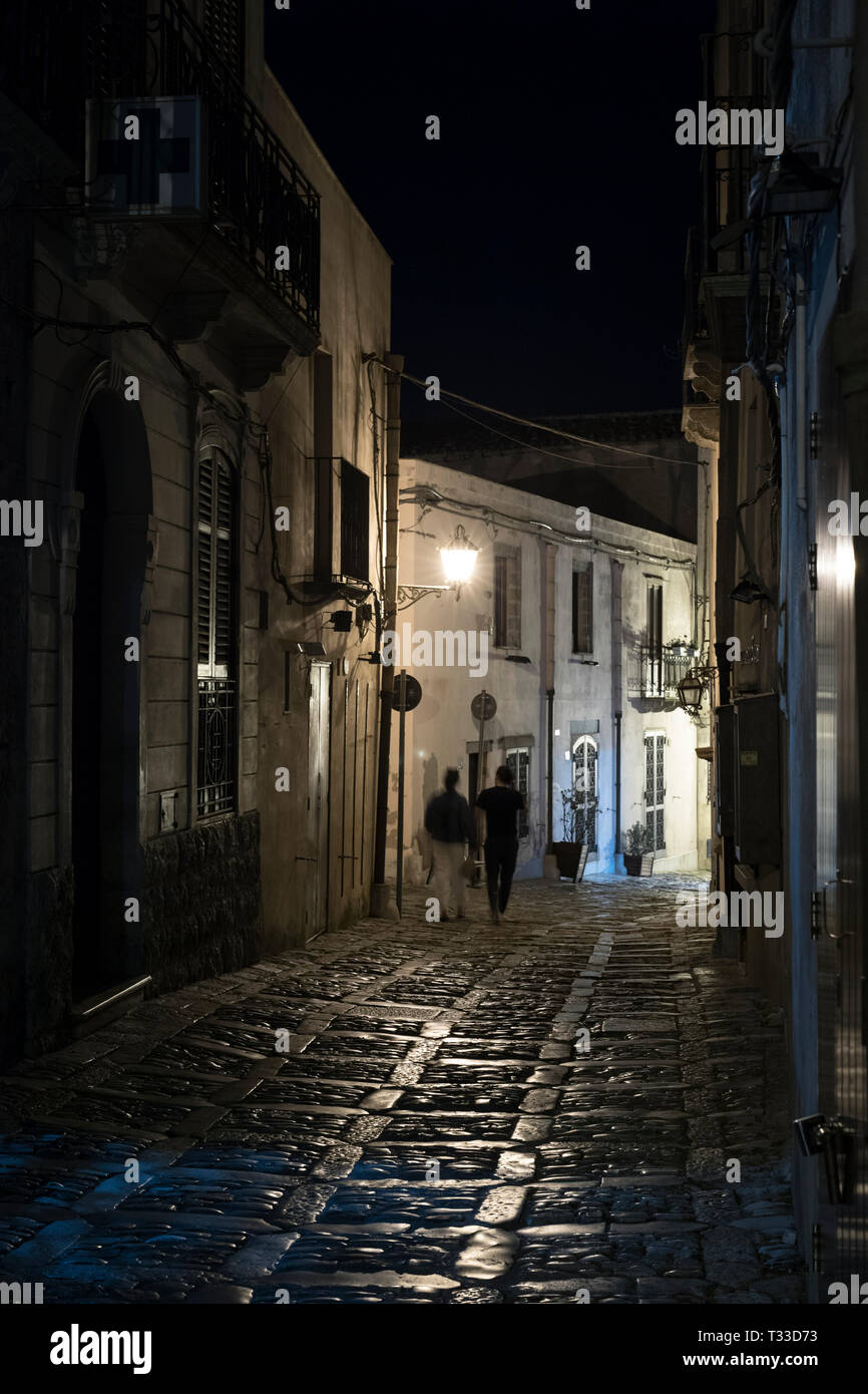 Lampade che brilla sulla pavimentazione di pietra di notte come turista giovane passeggiata lungo la strada di ciottoli vicolo di Erice, in Sicilia, Italia Foto Stock