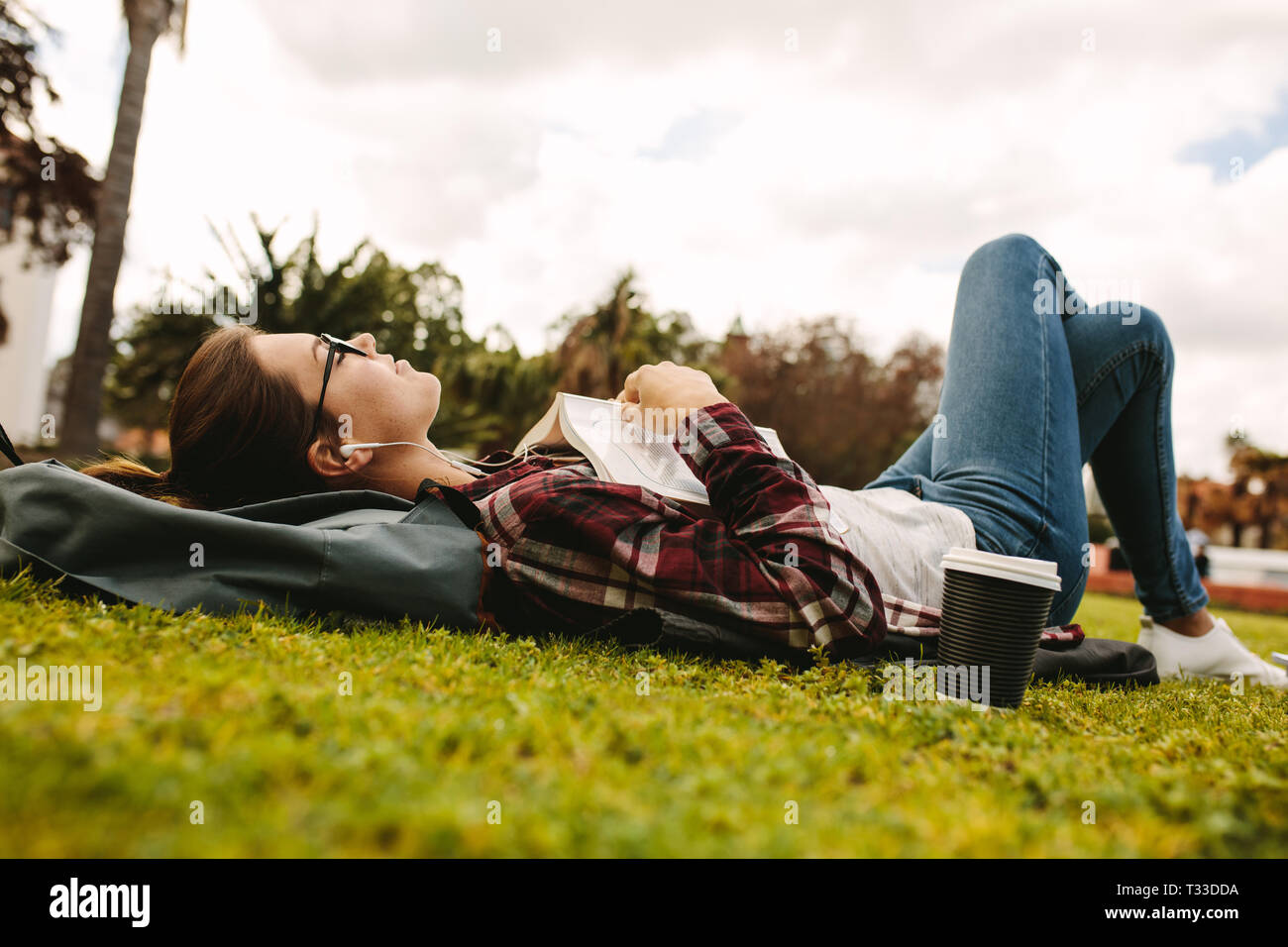 Donna studente con gli auricolari sdraiati sull'erba con libro. Ragazza di appoggio e di ascoltare musica durante la lettura del libro al campus universitario. Foto Stock