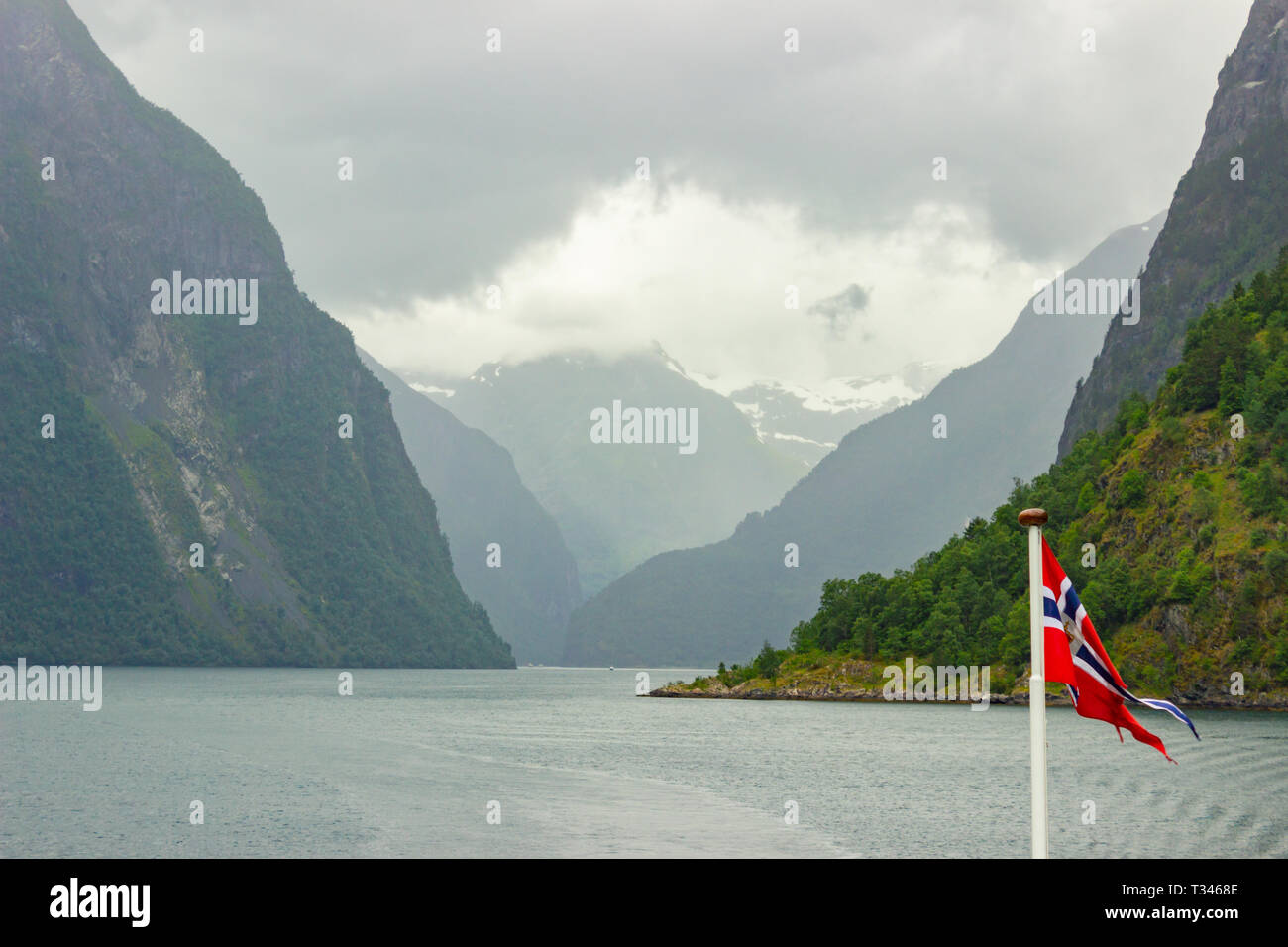 Bandiera della Norvegia sventolare nel vento su un traghetto che è voce lontano dalla terra. Nave scia del tracciato su acqua dietro un vaso, montagne distanti, bella evenin Foto Stock