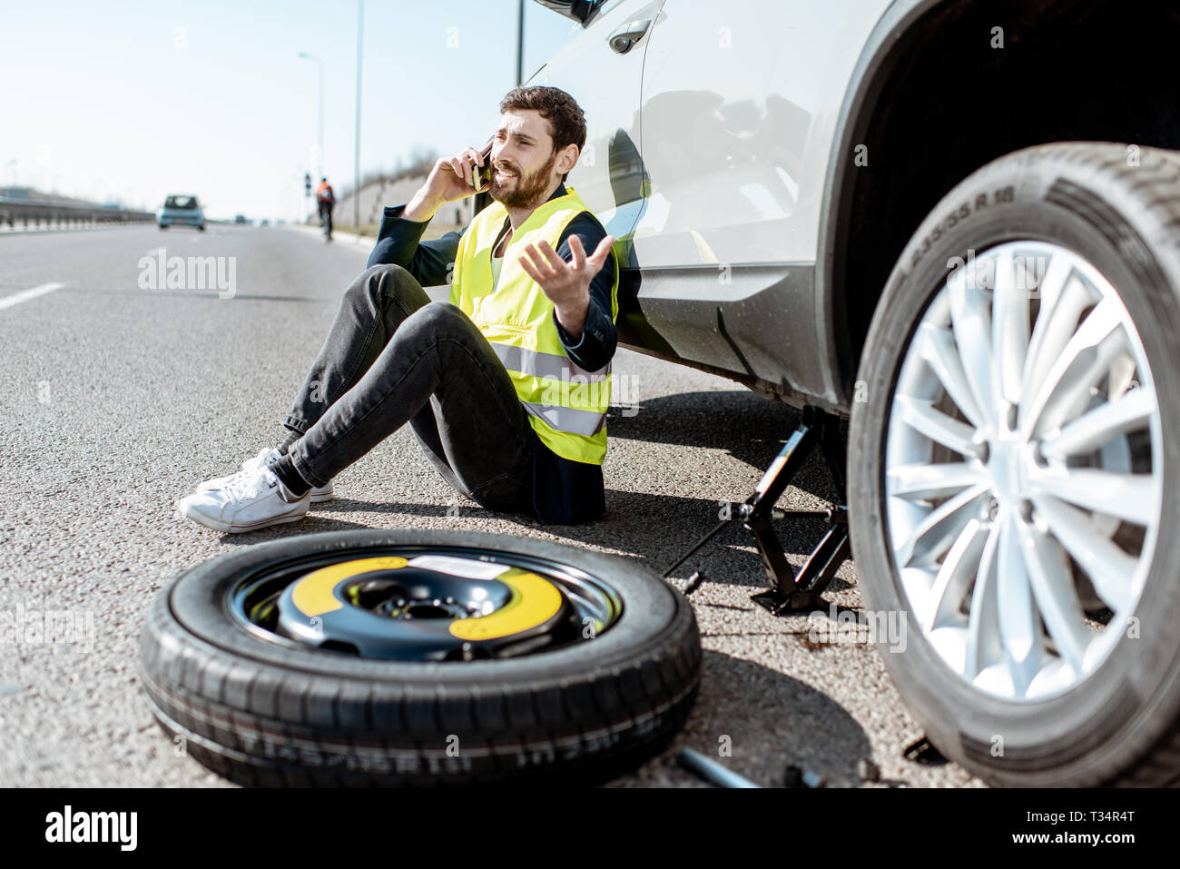 L'uomo chiamando l'assistenza stradale seduta vicino alla macchina per la rottura di una ruota sul ciglio della strada Foto Stock