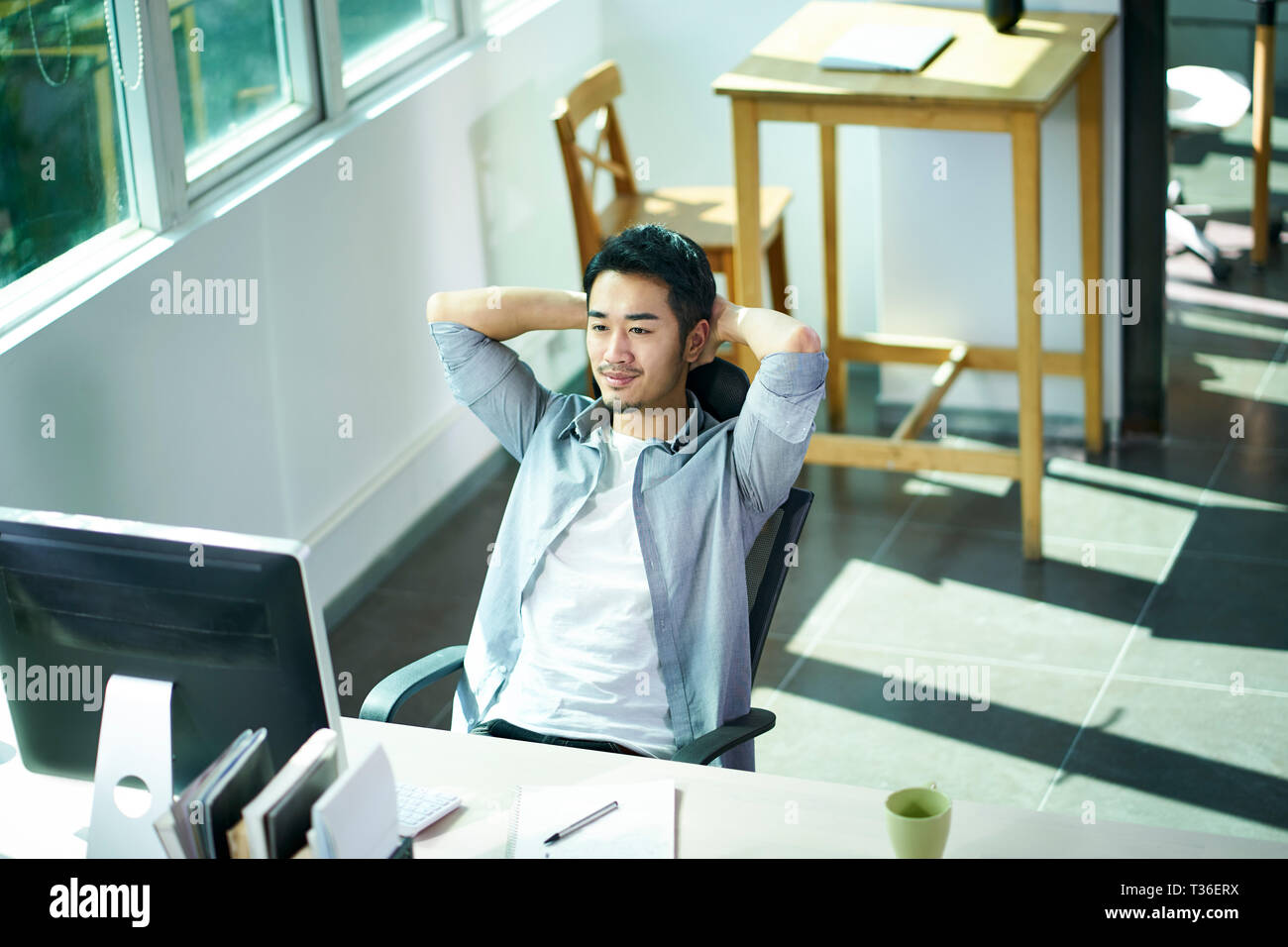 Giovani asiatici business man guardando il monitor di un computer con soddisfazione e con le mani dietro la testa. Foto Stock