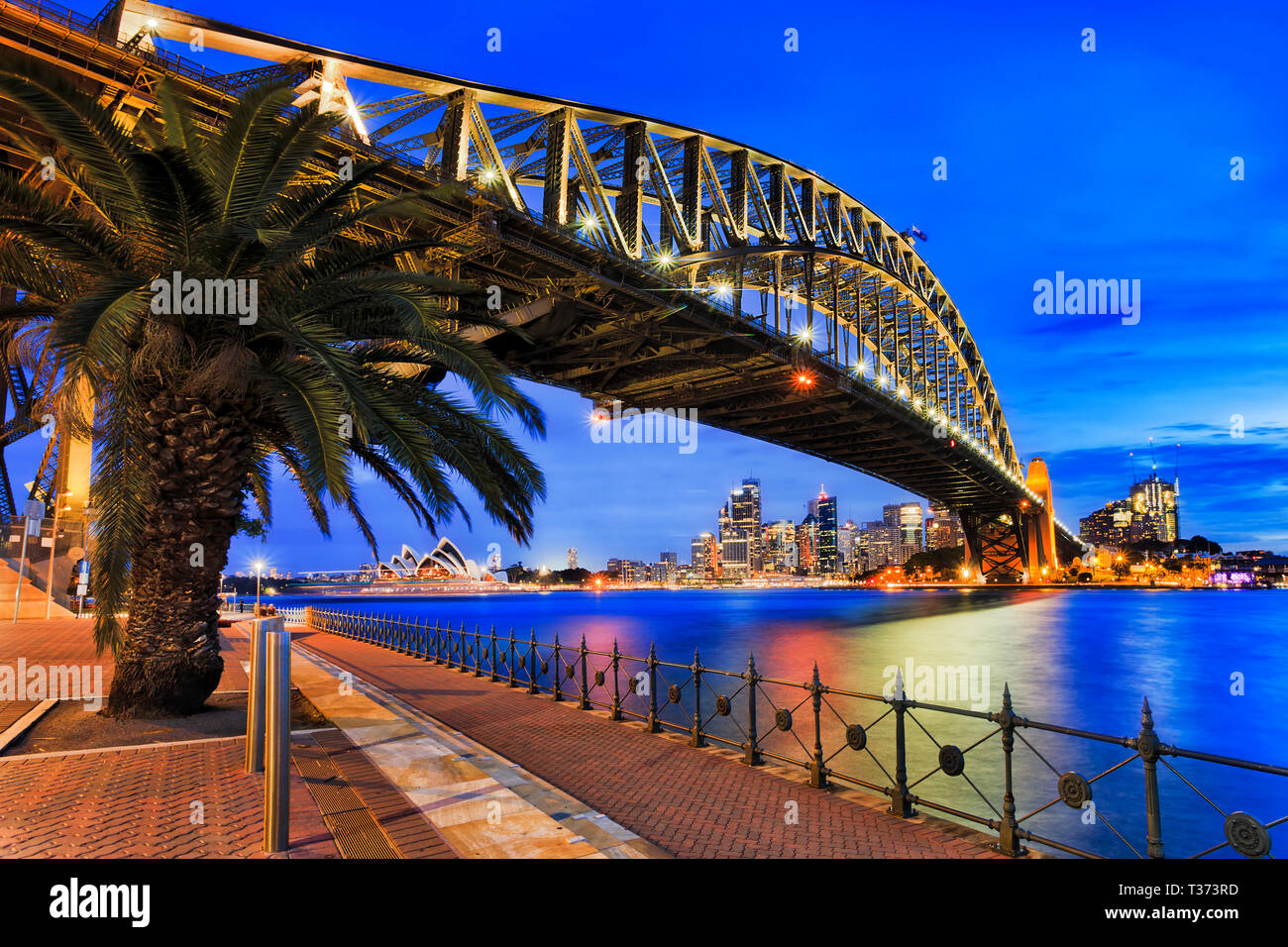 Vista laterale di acciaio arcata del ponte del Porto di Sydney durante il blu ora a Sunset over sfocato acque del porto di Sydney verso le rocce e il CBD di Città w Foto Stock