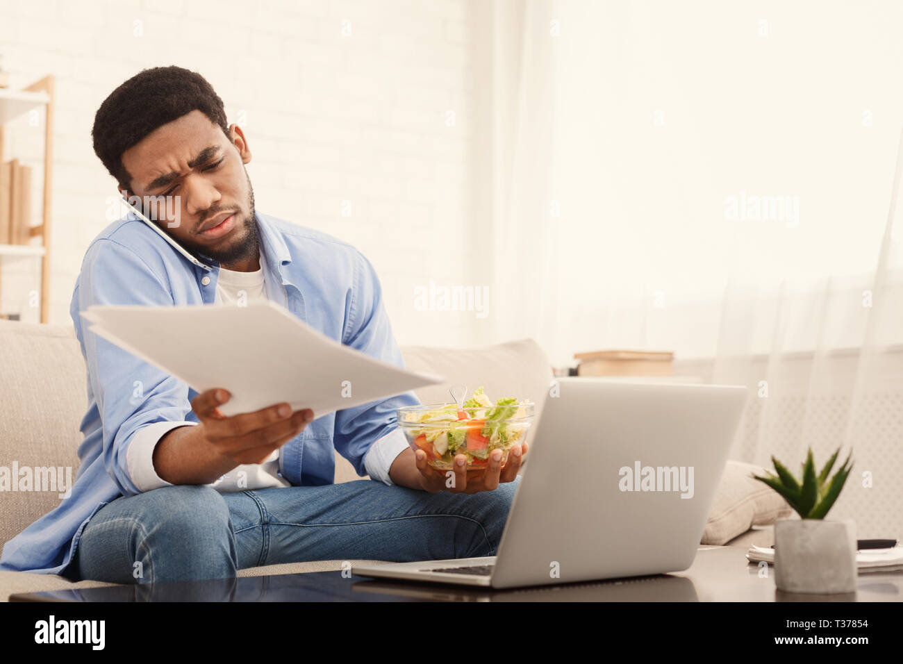 Non è tempo di pausa. Sistema nervoso african american guy mangiare sano colazione, parlando al telefono cellulare e di lavoro sul portatile a casa, spazio di copia Foto Stock
