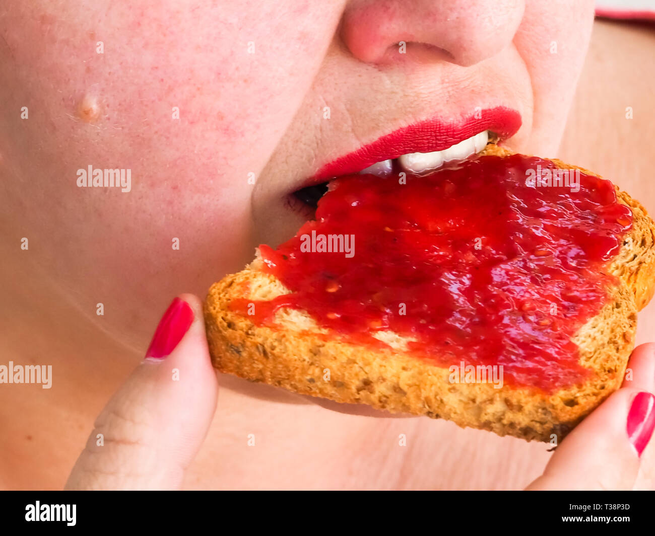 Una donna adulta con unghie rosse e le labbra di mangiare una fetta biscottata con confettura di lamponi Foto Stock