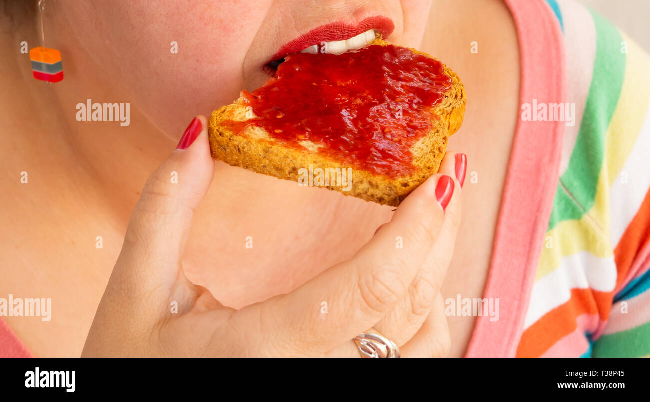 Una donna adulta con unghie rosse e le labbra di mangiare una fetta biscottata con confettura di lamponi Foto Stock