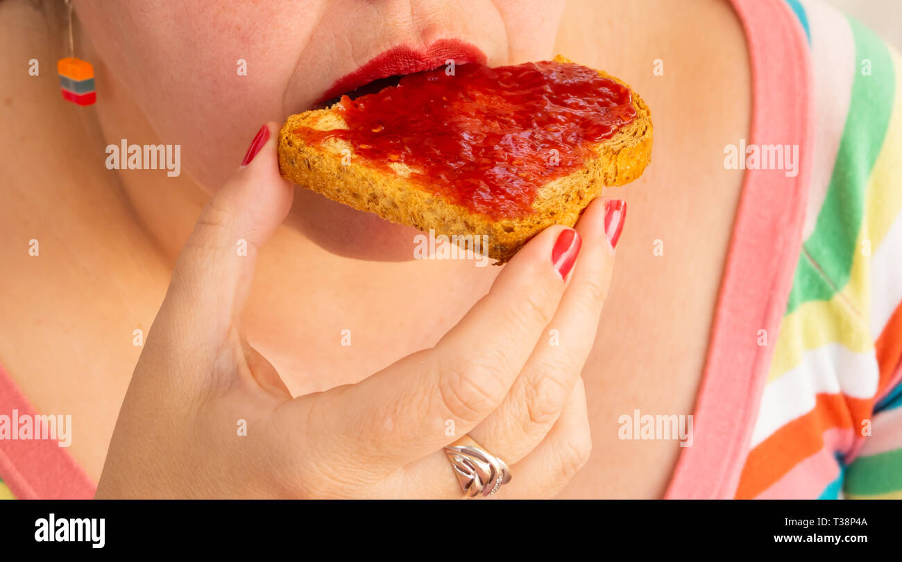 Una donna adulta con unghie rosse e le labbra di mangiare una fetta biscottata con confettura di lamponi Foto Stock