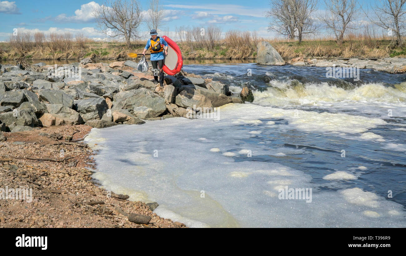 Senior paddler è gonfiabile portaging stand up paddleboard oltre un rapido - South Platte River in primavera scenario con una bassa portata d'acqua. Foto Stock