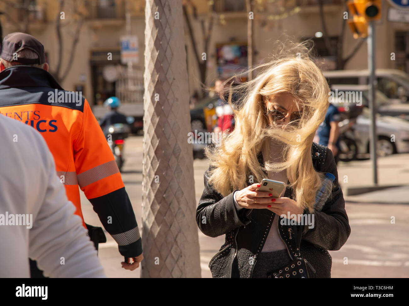 Spagna, Barcellona, 14 marzo 2019.Una ragazza con lunghi capelli biondi e occhiali scuri si erge nel mezzo di una strada trafficata e guarda al suo telefono.Il vento è Foto Stock