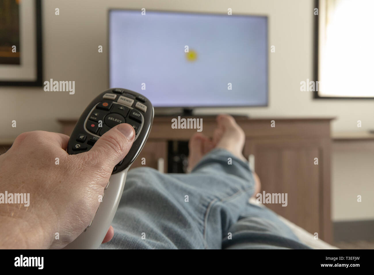 Mano che tiene il telecomando per la televisione durante i momenti di relax con i piedi puntellato Foto Stock