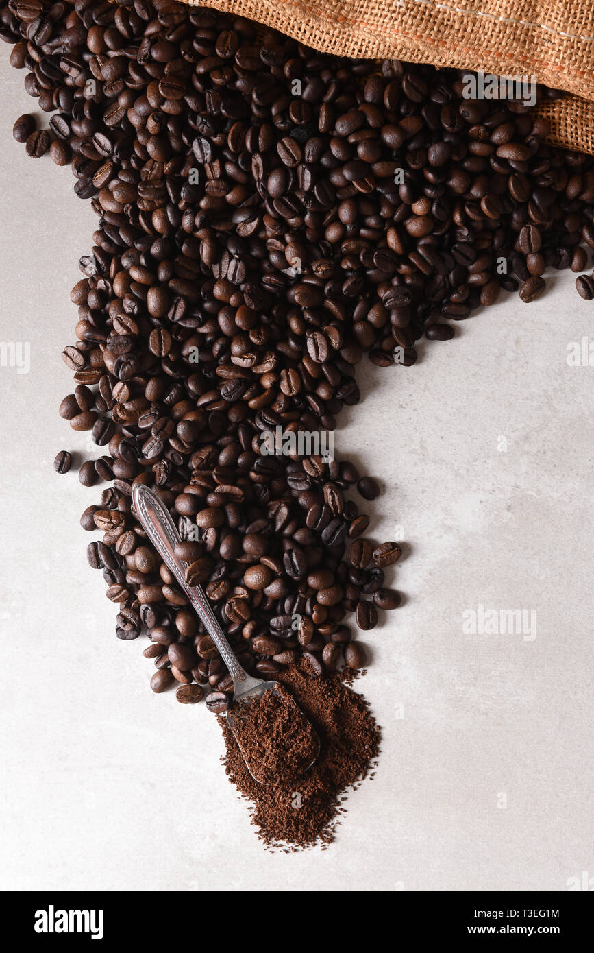 Elevato angolo di visione di un sacchetto di tela di caffè torrefatto in grani che si rovescia sulla superficie con un cucchiaio di caffè macinato al termine del versamento. Foto Stock