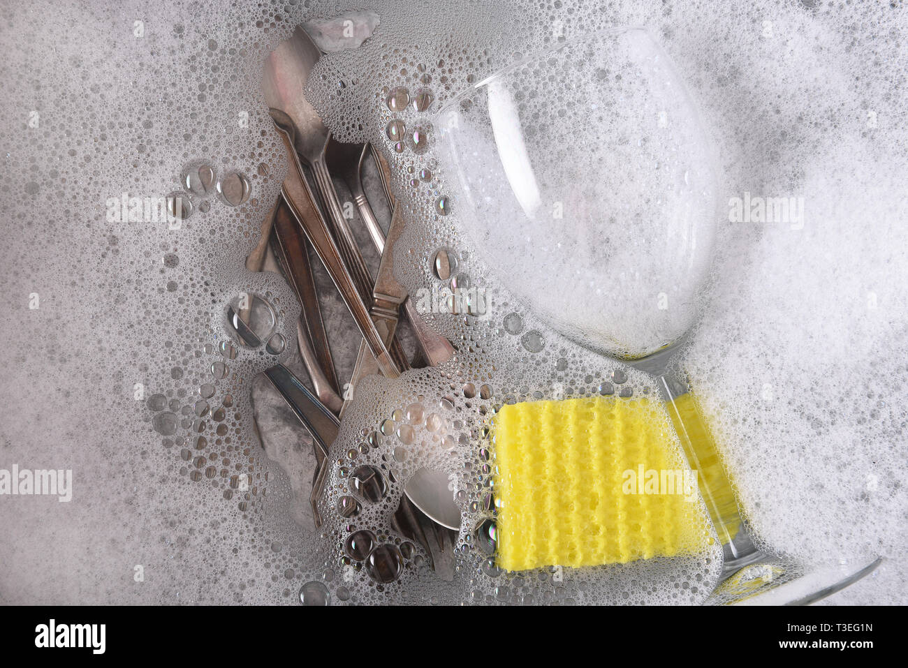 Scatto dall'alto di un dishpan riempito con acqua saponata con posate, un bicchiere di vino e una spugna. Foto Stock