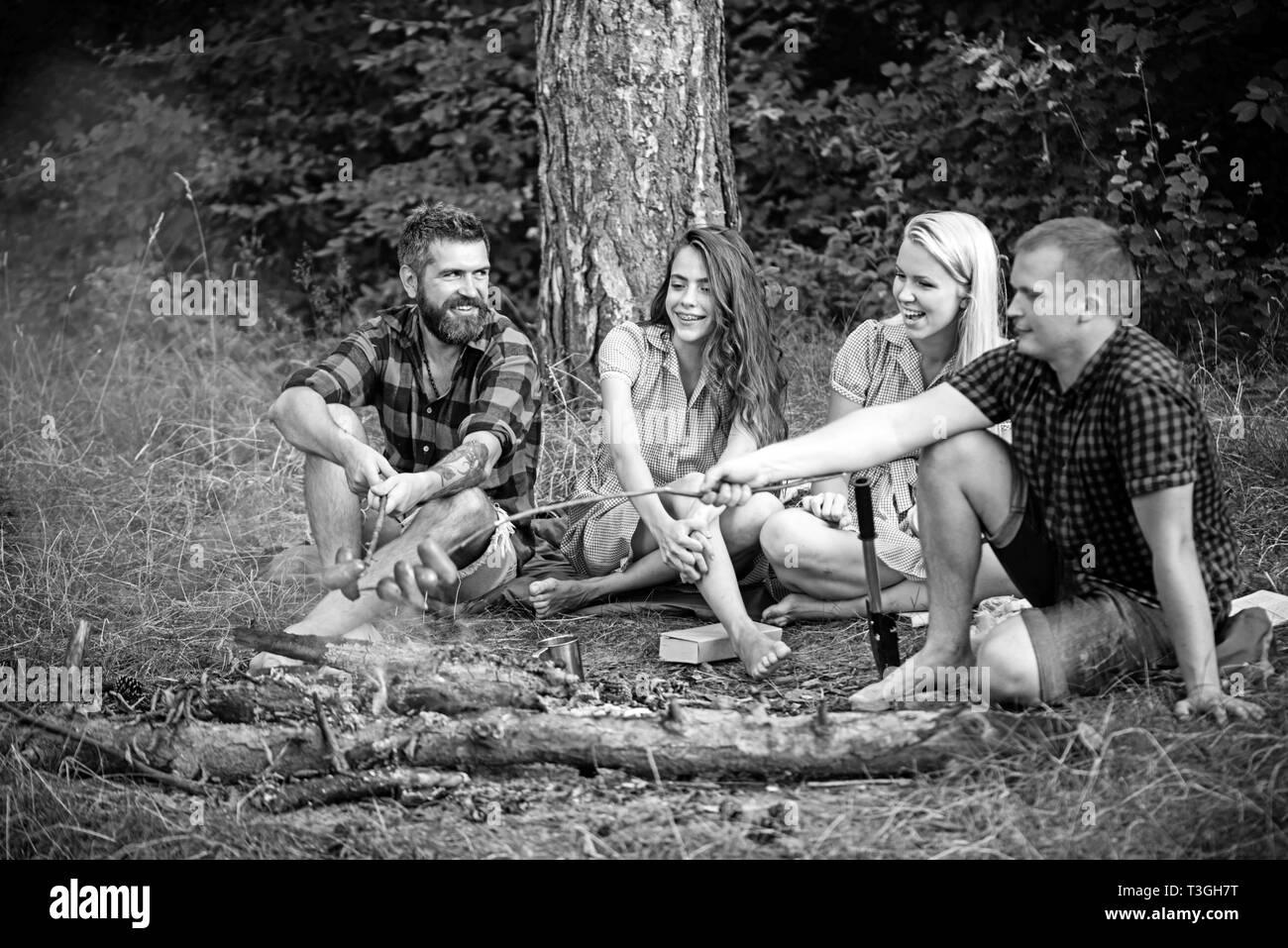 Coppie giovani avente picnic nei boschi. Uomo Barbuto e il suo migliore amico la frittura salsicce sul fuoco. Le persone felici seduti intorno al falò, amicizia e Foto Stock