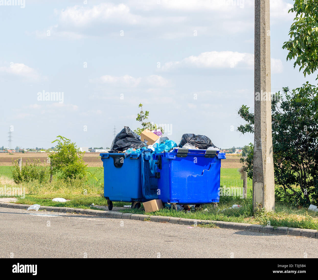 Traboccante di plastica blu lattine di rifiuti con il nero di sacchi, scatole di cartone e di altri rifiuti al di fuori della città vicino alla strada. Uno dei rifiuti può è rotto Foto Stock