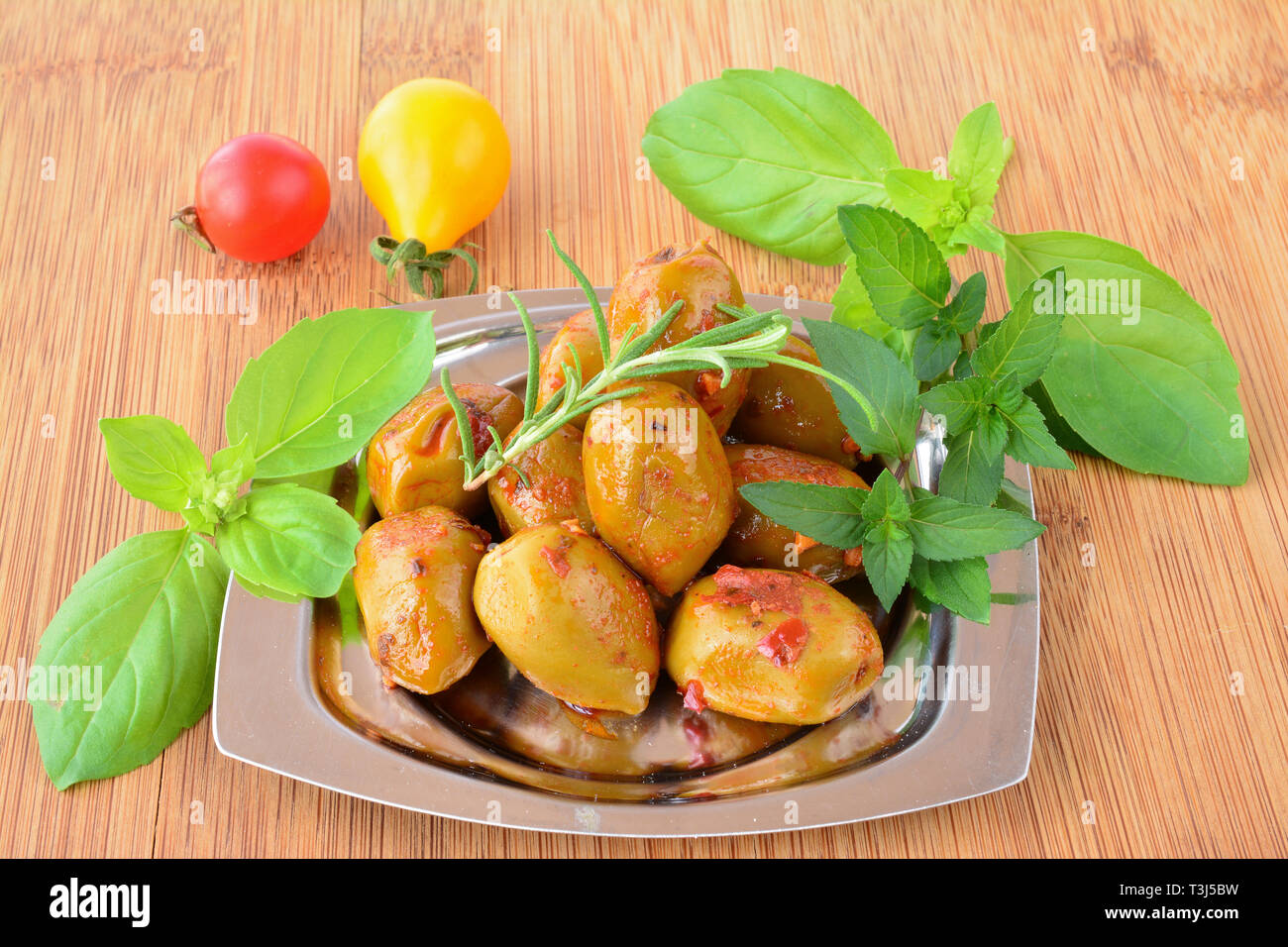 Piccante, hot olive verdi con fresche, verdi piante aromatiche e pomodoro ciliegino in piccole, metallo piattino in bambù tavolo in legno Foto Stock