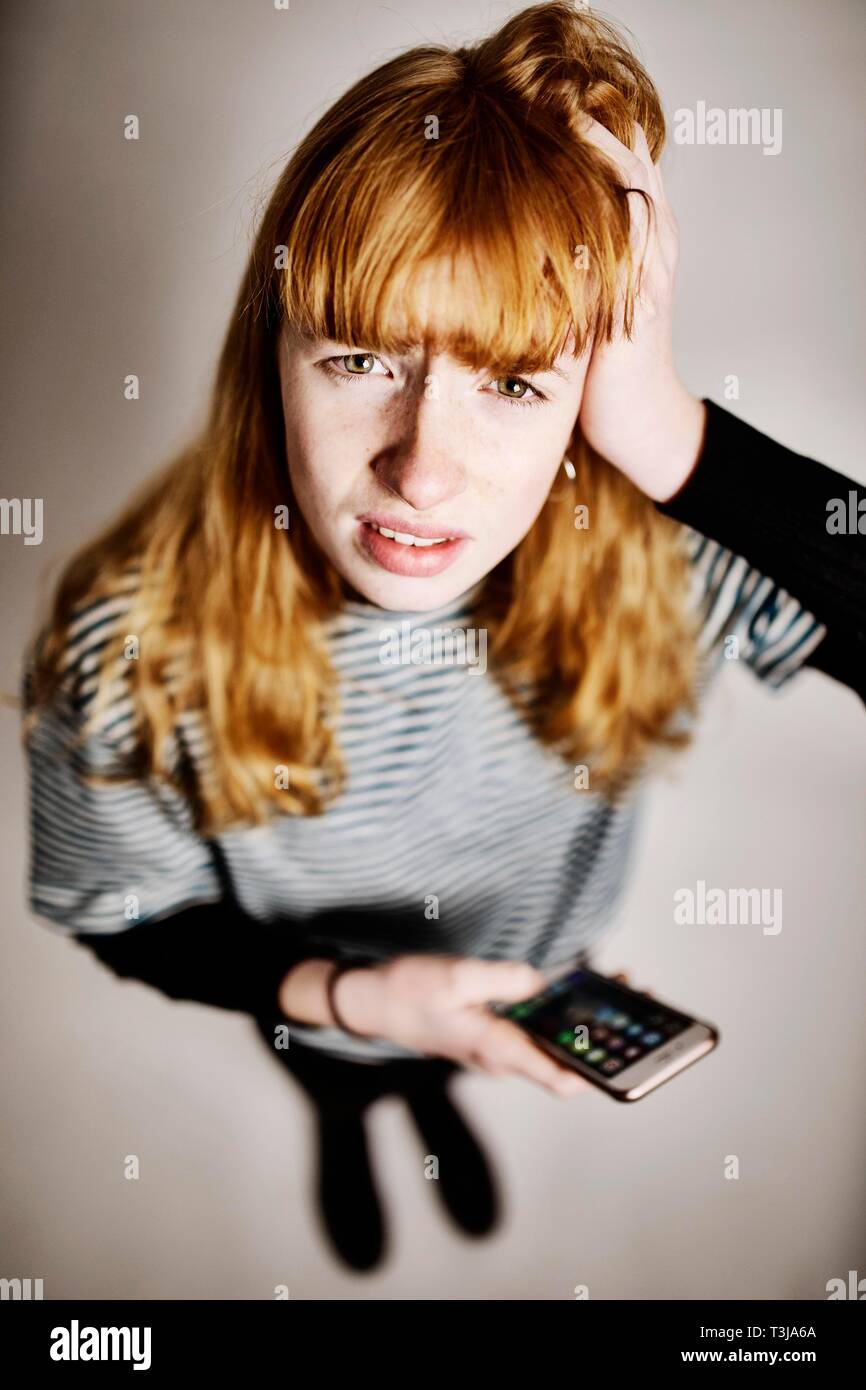 Ragazza adolescente, i capelli rossi, con lo smartphone in mano, disperatamente tirando i suoi capelli, studio shot, Germania Foto Stock
