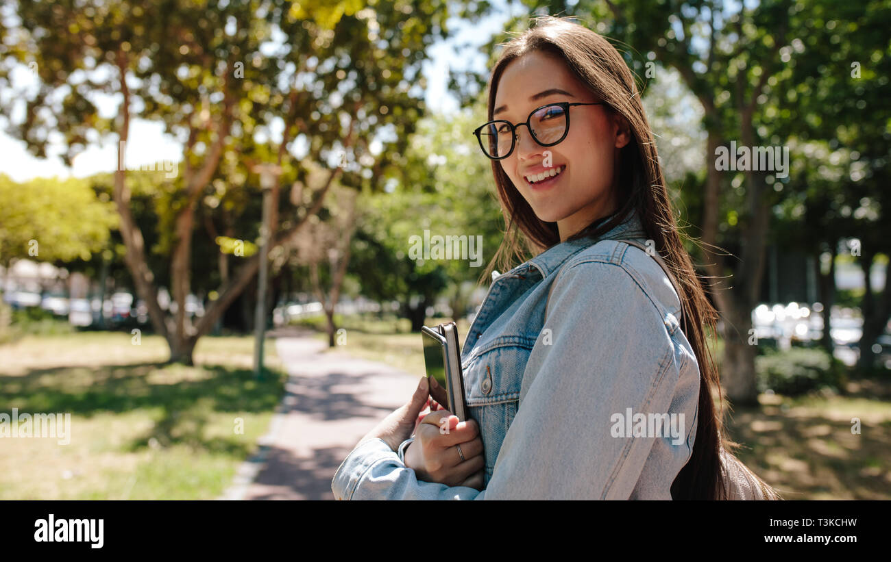 Vista laterale di un asiatico studente di college in piedi sul campus tenendo i suoi libri. Ragazza sorridente in occhiali in piedi in un parco in una giornata di sole. Foto Stock