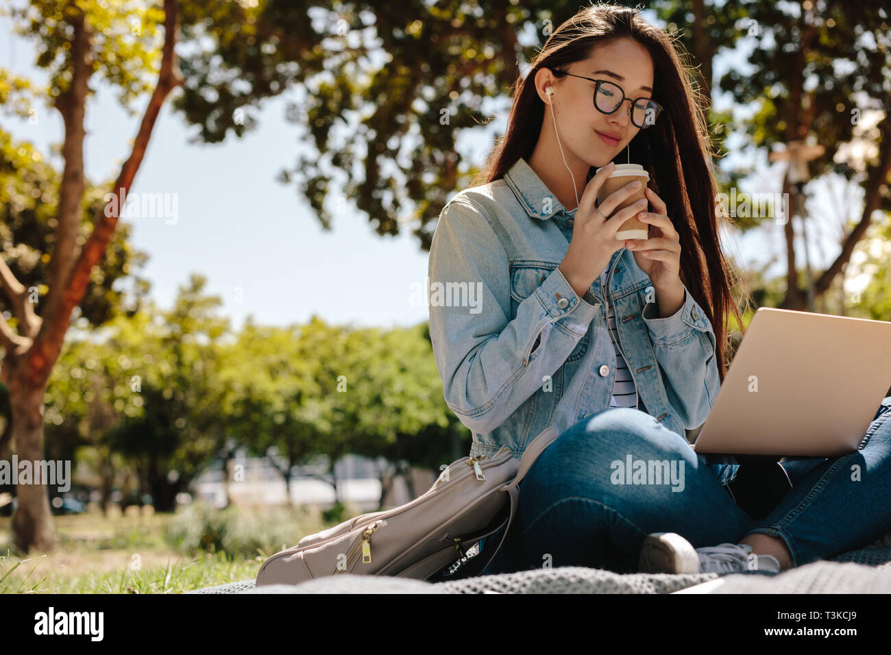 Allegro asian college ragazza seduta in un parco con un computer portatile mentre si gusta un caffè. Close up di un sorridente ragazza seduta all'aperto in un parco rel Foto Stock