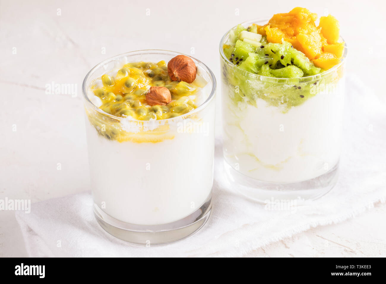 Close-up due bicchieri con il bianco yogurt fatto in casa e frutta fresca purea di frutto della passione, kiwi e mango sono in piedi sulla cucina bianca tovagliolo bianco su t Foto Stock