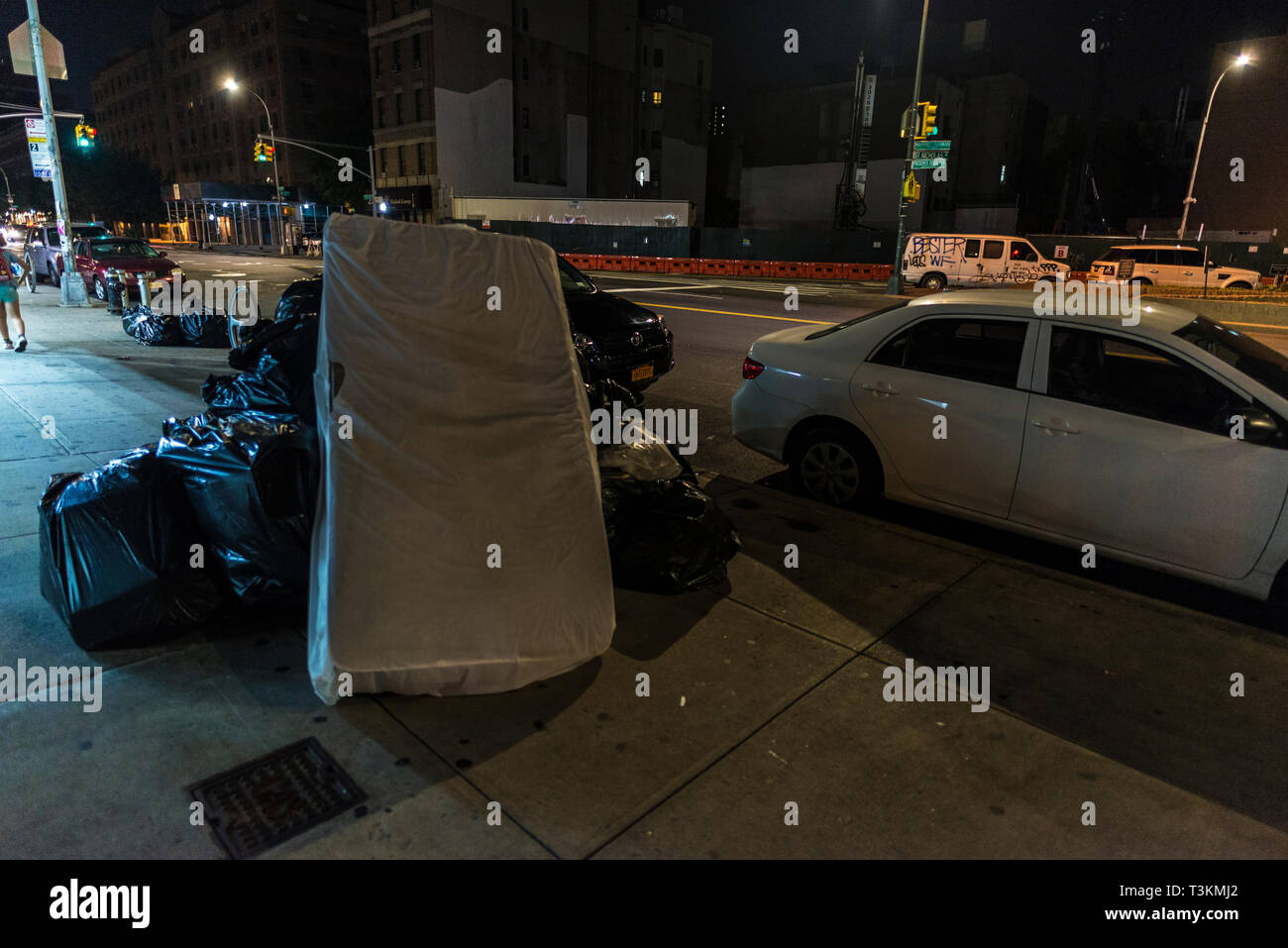 La città di New York, Stati Uniti d'America - 30 Luglio 2018: sacchetto di immondizia e un materasso su una strada di notte con la gente intorno in Harlem, Manhattan, New York City, Stati Uniti d'America Foto Stock