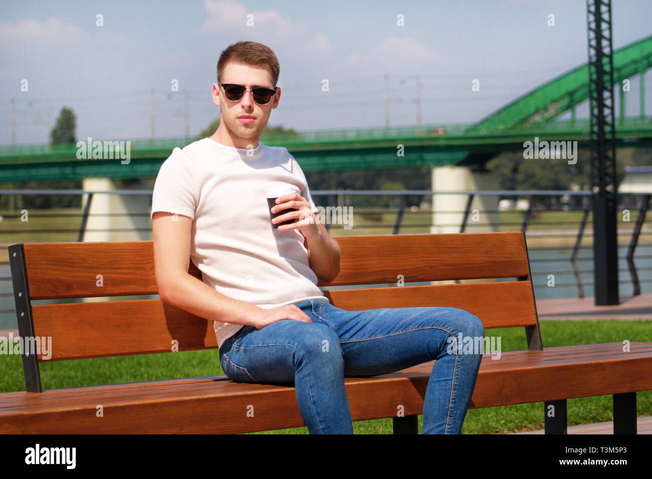 Bel ragazzo che indossa gli occhiali da sole, seduta su una panchina su un  molo e di bere il caffè per andare. Prendendo una pausa dalla città il  lettore RUSH. Lo sfondo