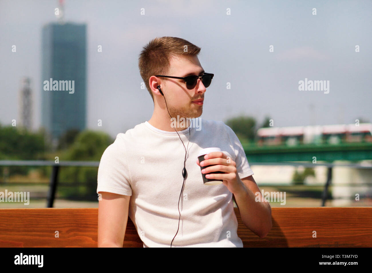 Bel ragazzo che indossa gli occhiali da sole con tablet e cuffie, seduta su  una panchina e di bere il caffè per andare. Bello studente immerso in  social media Foto stock -