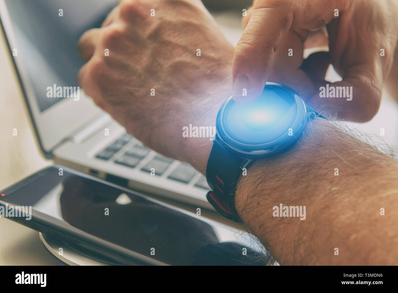 Uomo che utilizza smartwatch con il suo dito in ufficio, con lo smartphone e il computer portatile in background Foto Stock