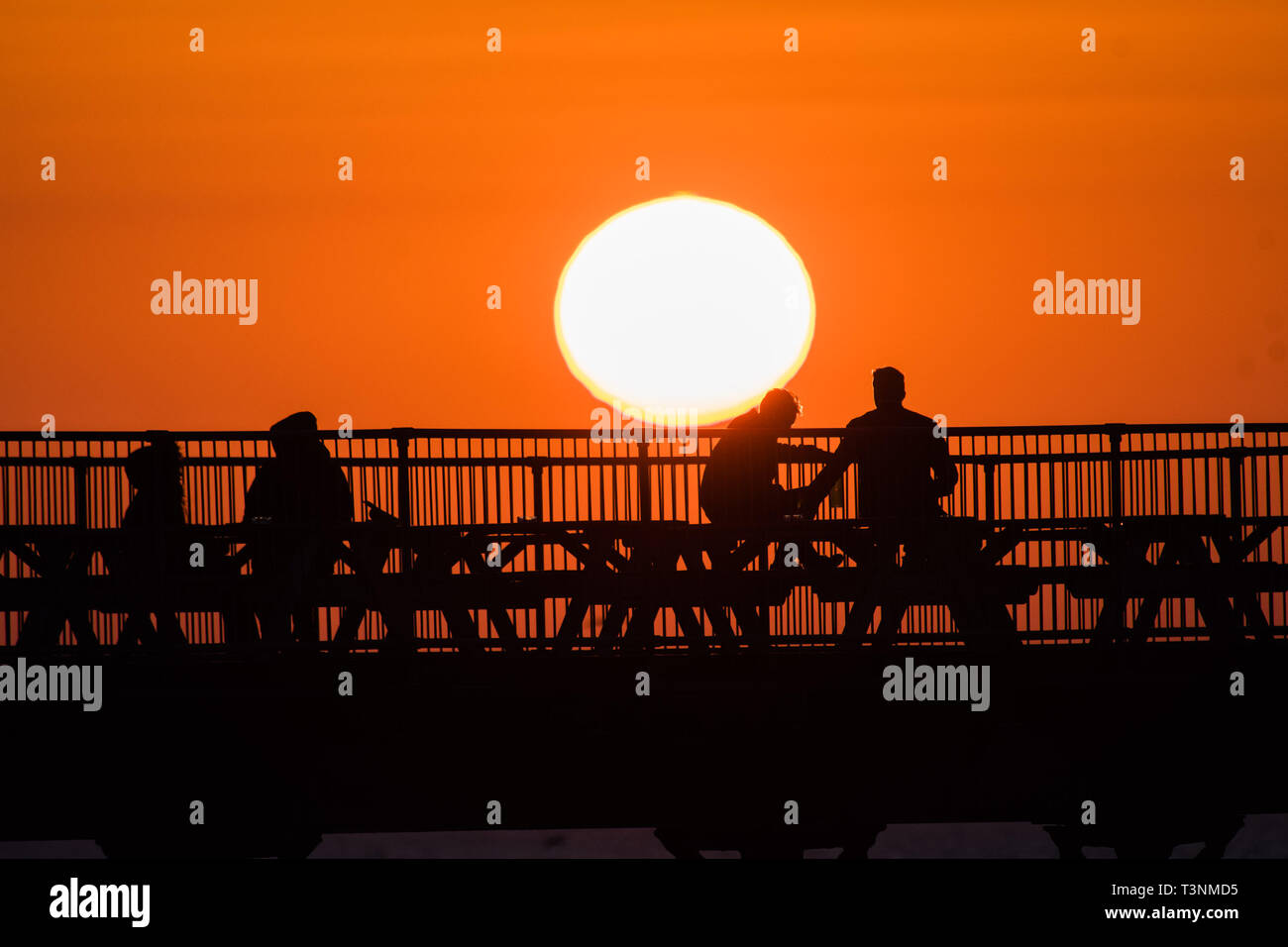 Aberystwyth, UK. Decimo Apr, 2019. Regno Unito: meteo alla fine di una giornata ininterrotte blu del cielo e il caldo sole primaverile, il sole tramonta spettacolarmente dietro le sagome delle persone gustando un drink sul mare dal molo di Aberystwyth su Cardigan Bay costa del Galles occidentale. Credito: keith morris/Alamy Live News Foto Stock