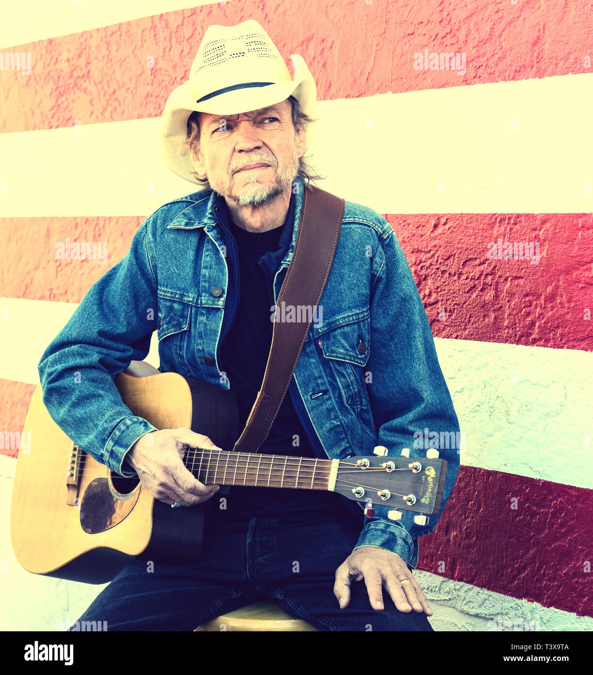Il vecchio cowboy con barba grigia riproduce una chitarra mentre in piedi accanto ad un bianco e rosso parete. Foto Stock
