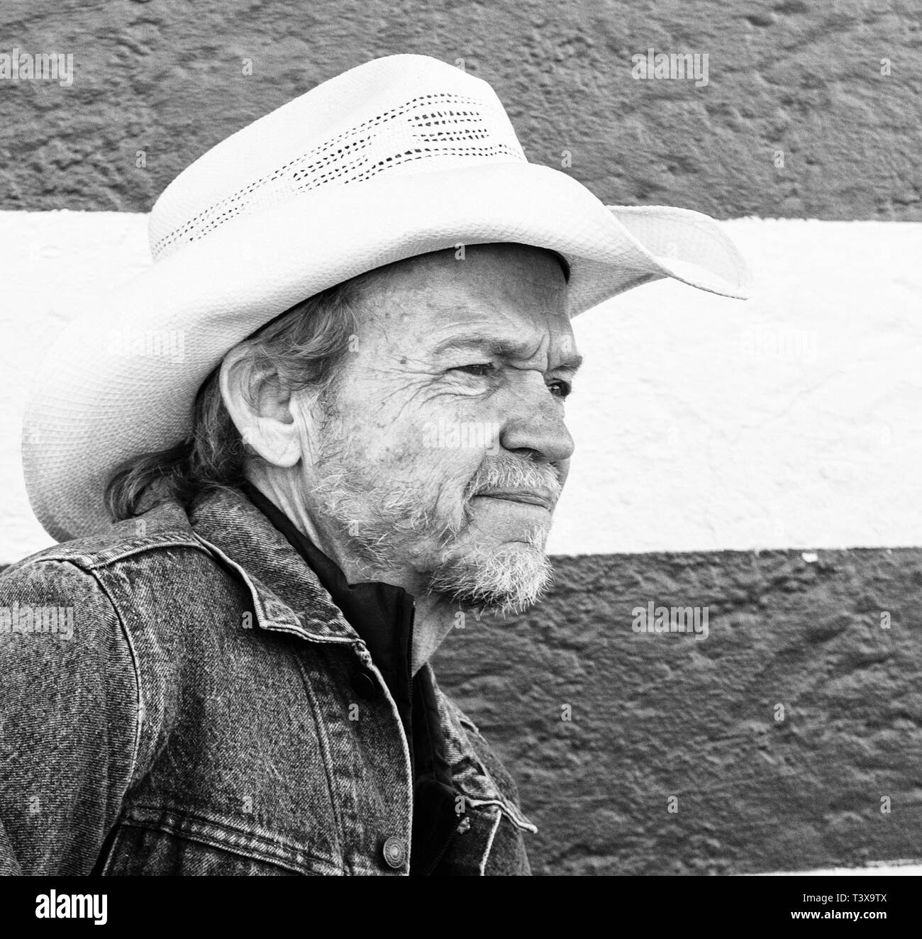 Il vecchio cowboy con cappello da cowboy e barba grigia appare su di un lato mentre in piedi accanto ad un bianco e nero parete a strisce. Foto Stock