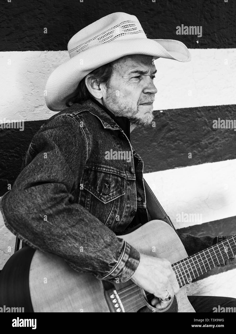 Il vecchio cowboy con cappello da cowboy e barba grigia riproduce una chitarra mentre in piedi accanto ad un bianco e nero parete a strisce. Foto Stock