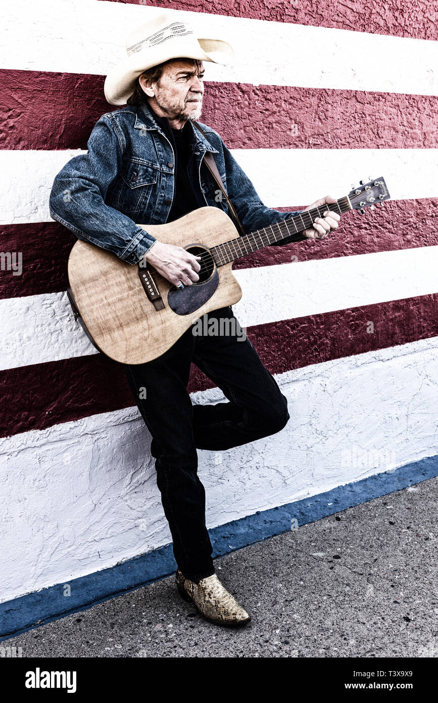 Il vecchio cowboy con barba grigia riproduce una chitarra mentre in piedi accanto ad un bianco e rosso parete. Foto Stock
