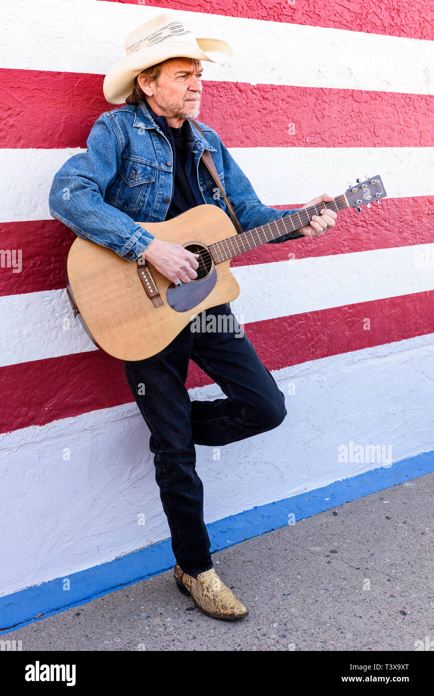 Il vecchio cowboy con barba grigia riproduce una chitarra mentre in piedi accanto ad un bianco e rosso parete. Foto Stock