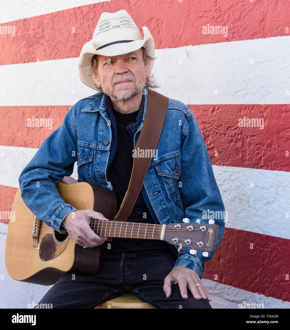 Il vecchio cowboy con barba grigia riproduce una chitarra mentre in piedi accanto ad un bianco e rosso parete. Foto Stock