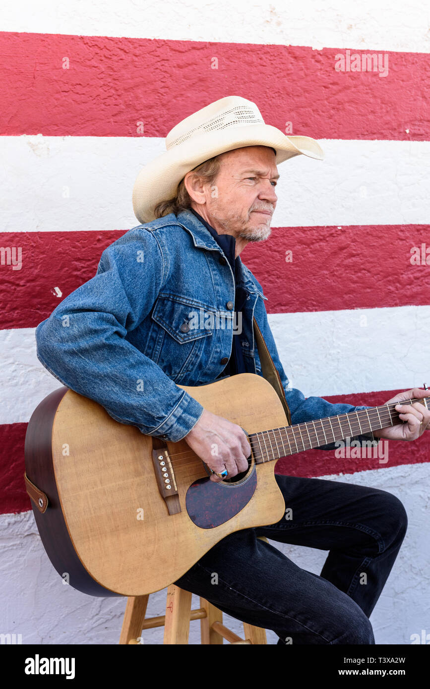 Il vecchio cowboy con barba grigia riproduce una chitarra mentre in piedi accanto ad un bianco e rosso parete. Foto Stock