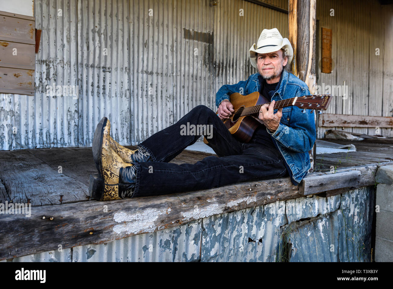 Il vecchio Cowboy indossa il cappello da cowboy e stivali riproduce una chitarra mentre è seduto su un vecchio dock. Foto Stock