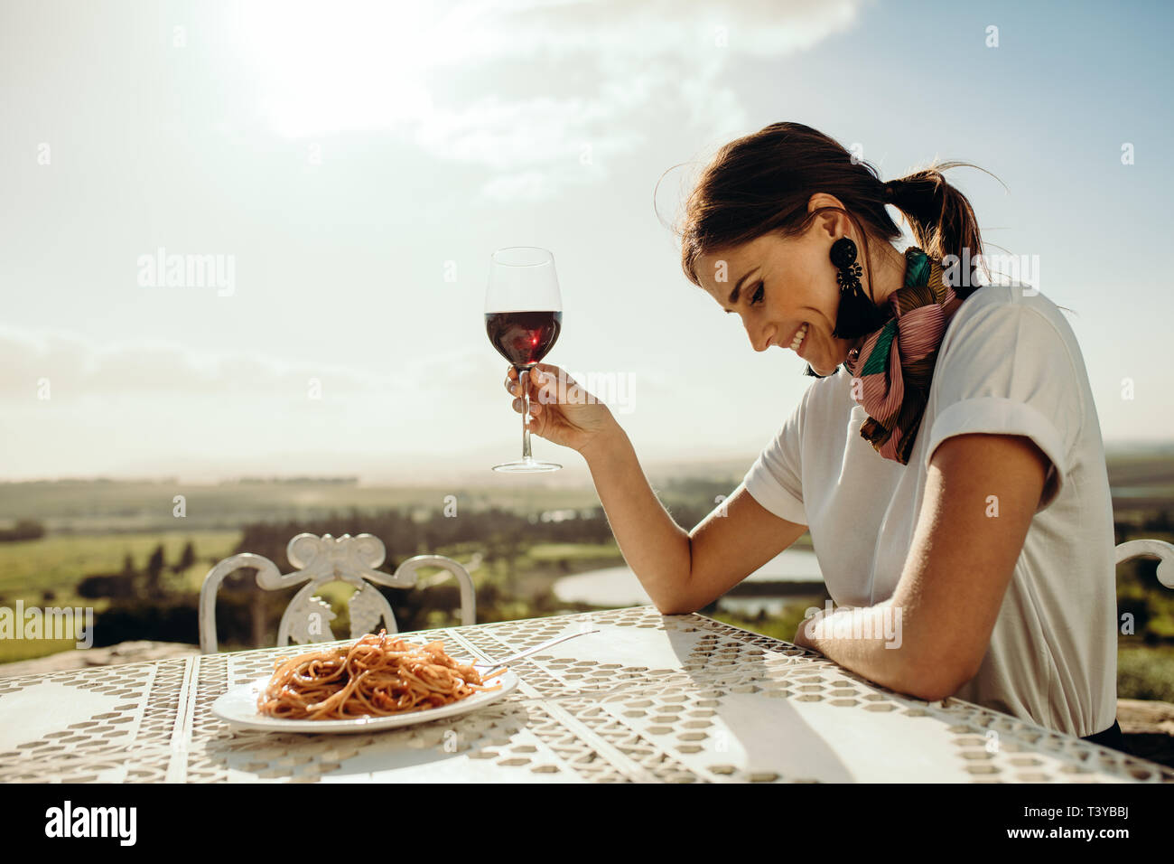 In prossimità di una donna per cenare fuori seduta a una tavola di ristorante. Vista laterale di un sorridente donna seduta al tavolo guardando in giù con un bicchiere di vino rosso Foto Stock
